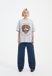 Světle šedé oversize tričko s motivem tygra a červeným textem, spárované s širokými džíny a černými špičatými botami.