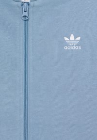 Veste zippée bleu clair en matériau doux, présentant un logo brodé blanc, une texture côtelée et une fermeture éclair à l'avant.