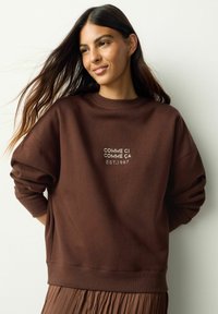 Braunes Sweatshirt mit entspannter Passform, ausgestattet mit einem gerippten Rundhalsausschnitt und einem gestickten Logodetail "COMME CI COMME ÇA EST.1987" auf der Vorderseite.
