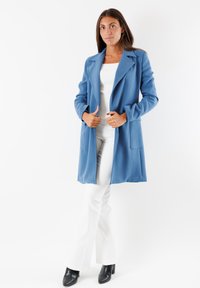 Cappotto azzurro chiaro a maniche lunghe con colletto a punte, tasche laterali e una texture liscia, indossato sopra una maglietta bianca e pantaloni bianchi.