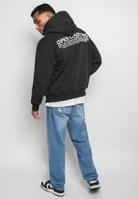 Schwarzer übergroßer Hoodie mit weißem Text auf der Rückseite, getragen über einem weißen Schichthemd, kombiniert mit hellblauen weiten Jeans und schwarzen Turnschuhen.