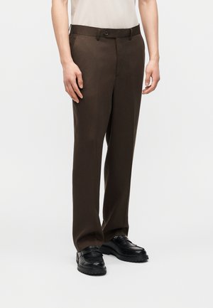 Vista de longitud de piernas de una persona que lleva pantalones marrón oscuro a medida, mocasines negros gruesos y una camisa blanca metida contra un fondo liso.