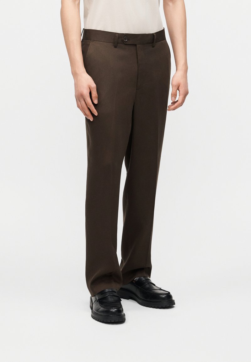 Άποψη μήκους ποδιών ενός ατόμου που φοράει σκούρο καφέ ραμμένο παντελόνι, μαύρα χοντρά loafers και ένα λευκό πουκάμισο μπα tucked μέσα σε απλό φόντο.