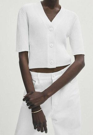 Personne portant un cardigan blanc côtelé à manches courtes boutonné et un pantalon blanc taille haute, les mains croisées devant.