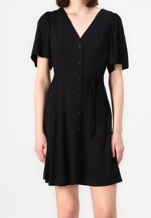 Femme portant une robe courte noire boutonnée avec un col en V, des manches évasées et une ceinture nouée sur le côté, debout devant un fond blanc uni.