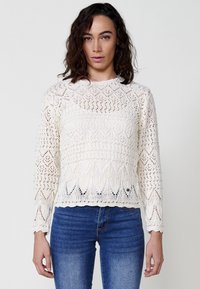 Suéter de punto color crema con intrincados patrones de diamante y hojas, mangas largas y dobladillo festoneado, combinado con jeans azules.