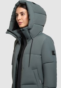 Chaqueta acolchada en gris apagado, que presenta una gran capucha, cierres de botón en la parte delantera, bolsillos laterales y una tela texturizada con un brillo sutil.