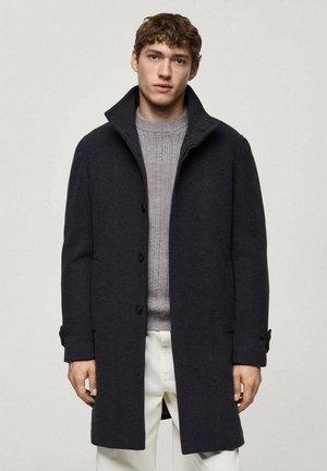 Dark wool coat with a high collar, featuring button closures and side pockets, worn over a light gray knit sweater and cream pants.