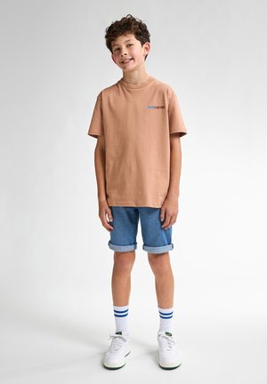 Camiseta de algodón color marrón claro con mangas cortas, con un logo en azul y marrón. Combinada con pantalones cortos de denim enrollados y zapatillas blancas.
