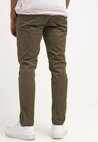 Chinos color khaki con una finitura in tessuto liscia, taglio slim e dettagli discreti sulle tasche posteriori, abbinati a sneakers bianche.