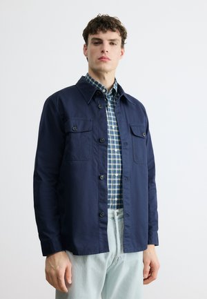 Joven con una sobrecamisa abotonada azul marino sobre una camisa de cuadros azul y blanca y pantalones azul claro, de pie frente a un fondo liso.