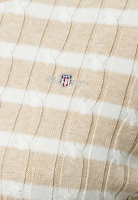 Beige und weiße gestreifte gestrickte Pullover mit strukturiertem Kabeldesign und gesticktem Logo auf der Brust. Weicher Stoff mit Ripp-Details.