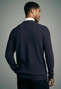 Strikket sweater i mørk lilla med et struktureret mønster, ribbede ærmer og kant. Båret over en hvid skjorte med krave, set fra bagenden.