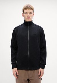 Giacca nera con zip in tessuto testurizzato, caratterizzata da un motivo geometrico, colletto alto e tasche laterali, indossata con pantaloni marrone chiaro.
