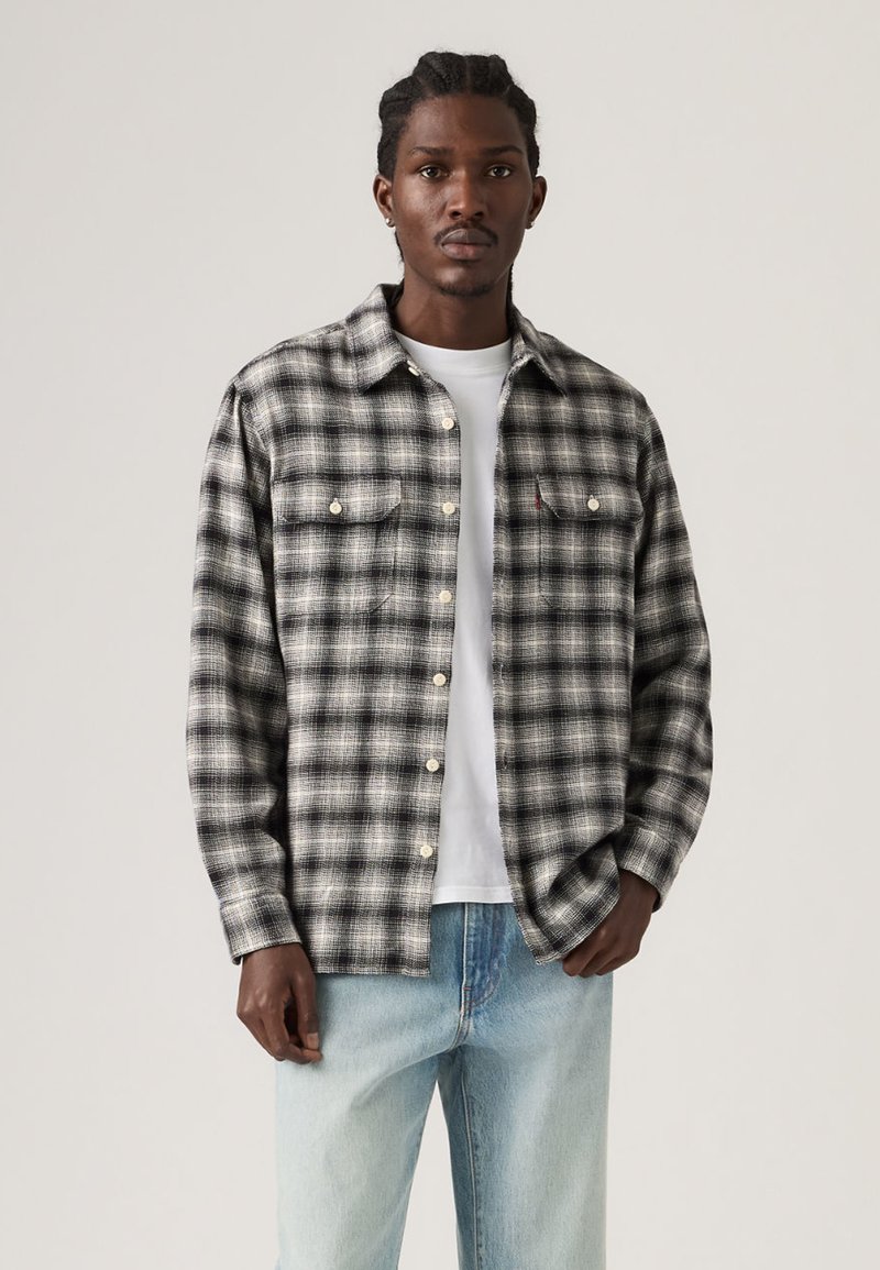 Homme portant une chemise à carreaux noir et blanc boutonnée par-dessus un t-shirt blanc et un jean bleu clair, debout devant un fond uni.