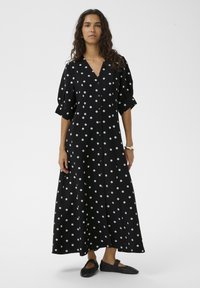 Fekete pöttyös maxi ruha V-nyakkivágással, gombos elülső résszel és rövid, fodros ujjakkal. Az anyag könnyűnek tűnik; légiesen áramló sziluettet mutat.