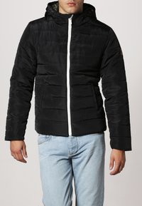 Sort pufferjakke med vandret quiltning, hvid lynlås og justerbar hætte. Båret over lyseblå jeans. Blød, glat stoftekstur.