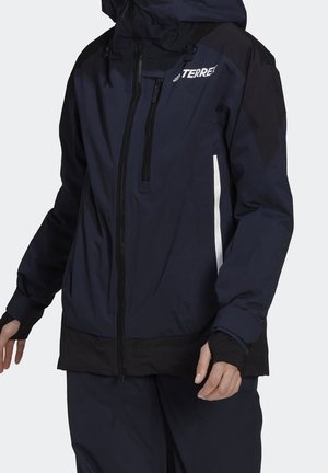 Person iført navyblå og sort Adidas Terrex outdoorjakke med hætte, lynlåslomme på brystet og tommelfingerhuller på ærmerne.