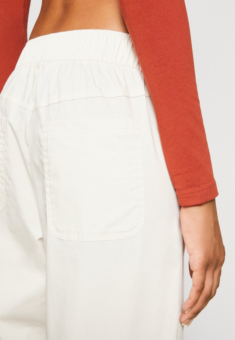 Weiße, elastische Bundhose mit Seitentaschen, aus strukturiertem Stoff; kombiniert mit einem eng sitzenden, langärmeligen Oberteil in rostfarbener Farbe.