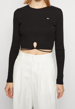 Femme portant un crop top noir à manches longues avec une découpe en forme de trou de serrure devant et un pantalon blanc taille haute sur un fond uni.