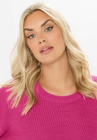 Fuksjowy sweter dzianinowy z okrągłym dekoltem i fakturowanym wzorem, z długimi włosami otaczającymi twarz z neutralnym makijażem.