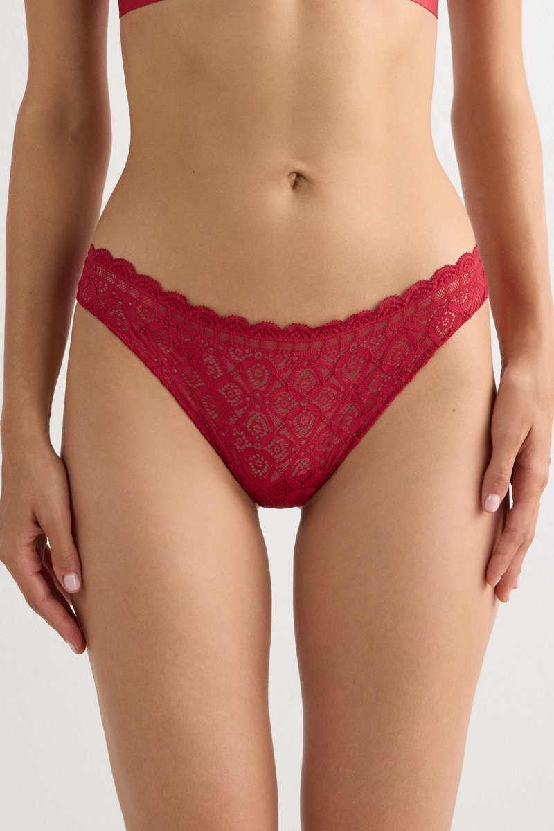 Buste féminin portant un sous-vêtement style bikini en dentelle rouge et un soutien-gorge assorti, debout avec les bras détendus le long du corps sur un fond uni.