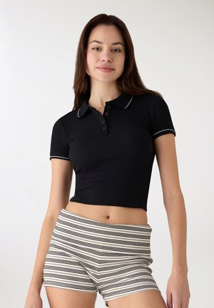 Polo nera a coste con collo a polo e cinque bottoni, rifinita con cuciture bianche. Abbinata a pantaloncini a righe crema e nere.