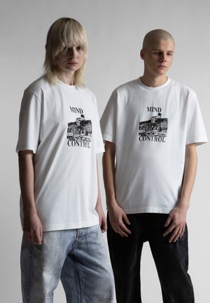 Dvaja mladí dospelí stoja vedľa seba, oblečení v bielych nadrozmerných tričkách s čiernym grafickým textom "MIND CONTROL" a vybledlých džínsach a čiernych nohaviciach.