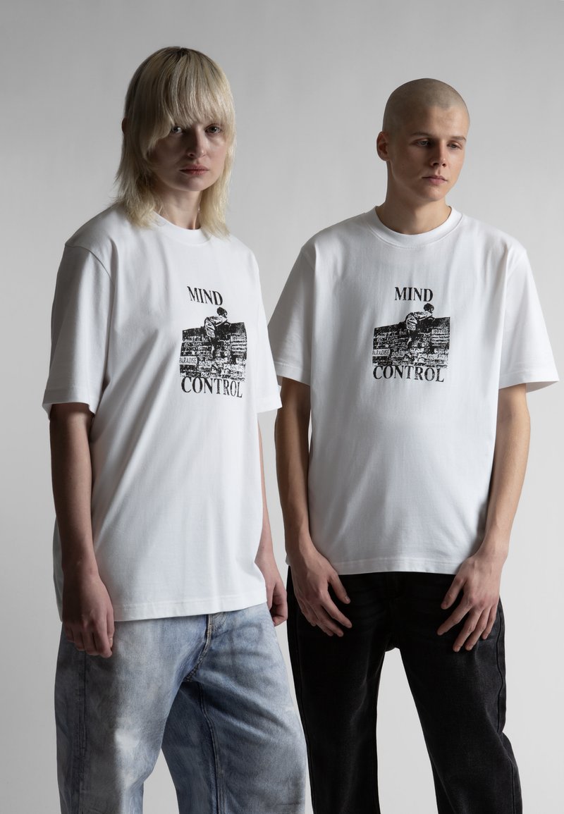 Δύο νεαροί ενήλικες στέκονται δίπλα δίπλα, φορώντας λευκά φαρδιά μπλουζάκια με μαύρο γραφικό κείμενο "MIND CONTROL" και ξεθωριασμένο τζιν και μαύρο παντελόνι.