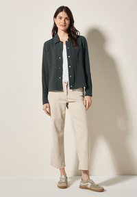 Camicia button-up verde scuro in tessuto texturizzato, abbinata a una canotta bianca e pantaloni beige chiaro, completata da sneakers grigie e bianche.