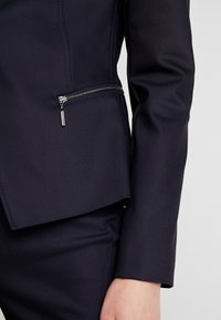 Giacca navy su misura realizzata in tessuto liscio, con tasca zip laterale e design aderente con maniche lunghe.