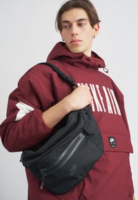 Mala tiracolo preta da Nike com bolsos com fecho, textura suave, apresentando um logotipo e detalhes em cinza. Usada sobre uma jaqueta bordo com capuz.