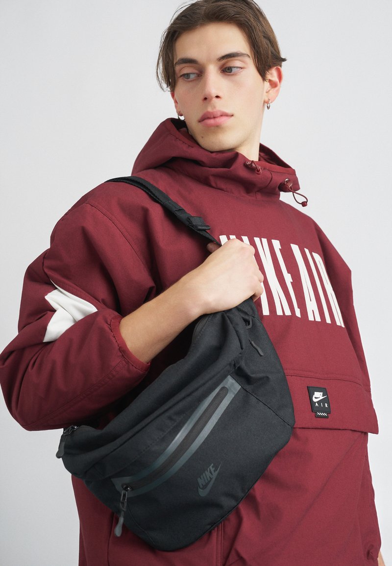 Mala tiracolo preta da Nike com bolsos com fecho, textura suave, apresentando um logotipo e detalhes em cinza. Usada sobre uma jaqueta bordo com capuz.