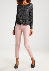 Kobieta w czarnej bluzce z długim rękawem w białe kropki i koronkowym wykończeniu, zestawiona z jasnoróżowymi dopasowanymi spodniami i czarnymi szpilkami.