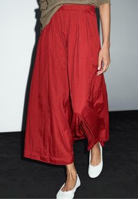 Punainen, poimutettu midihame, jossa on tekstuuri ja kiiltävä pinta. Yhdistetty valkoisten, litteiden kenkien kanssa. Vyötärö on joustava mukavuutta varten.