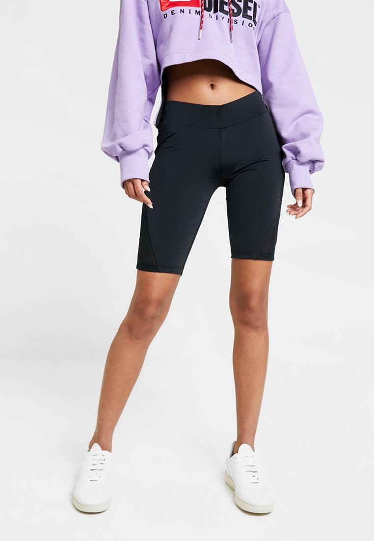 Sorte cykelshorts lavet af strækbart stof med en høj talje. Båret med en cropped lavendelfarvet sweatshirt med et logo. Hvide sneakers.