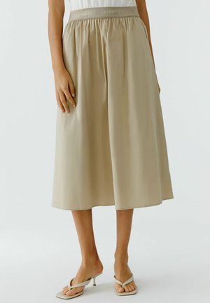 Frau trägt beige Culottes mit weitem Bein und elastischem Bund sowie beige Sandaletten mit Absatz, steht vor einem einfarbigen hellen Hintergrund.