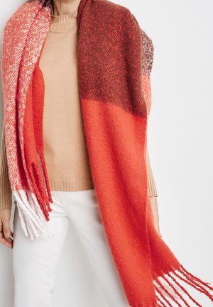 Femme portant un pull beige et un pantalon blanc, drapée d'une grande écharpe en laine à franges, dégradé de rouge et rose.