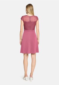 Robe rose avec un corsage transparent scintillant et une jupe évasée. Tissu lisse avec une taille marquée. Chaussures blanches à talons carrés.
