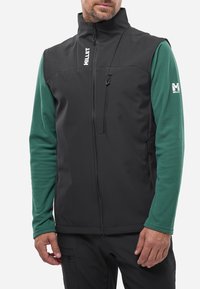 Schwarze Softshell-Weste mit hohem Kragen, Reißverschluss-Fronttasche und grünem Langarmshirt darunter. Mit Logo-Akzenten.