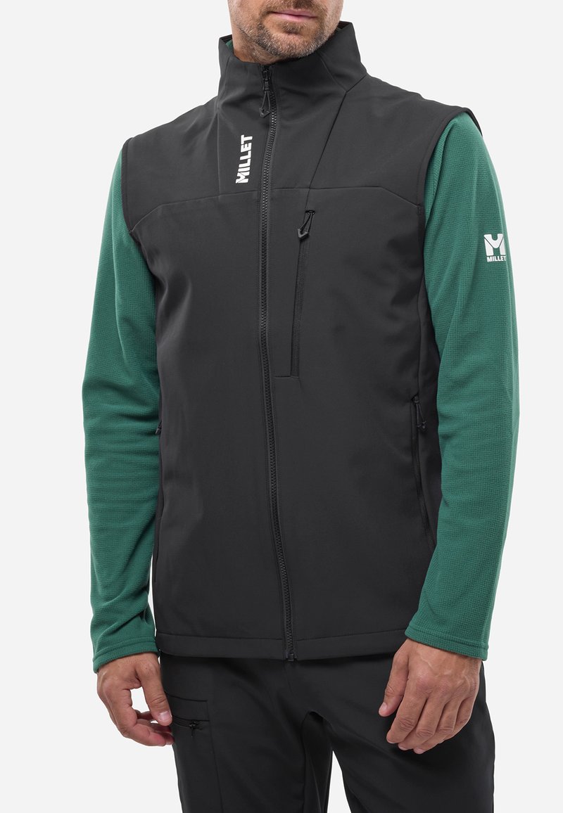Schwarze Softshell-Weste mit hohem Kragen, Reißverschluss-Fronttasche und grünem Langarmshirt darunter. Mit Logo-Akzenten.