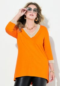 Orange Tunika mit V-Ausschnitt und Spitzenrand am Ausschnitt und an den Ärmeln, kombiniert mit schwarzen Leggings. Glatte Stoffqualität, lockere Passform.