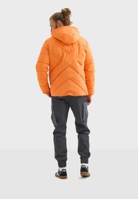 Chaqueta acolchada naranja con costuras en forma de chevrón, combinada con pantalones cargo gris oscuro que tienen bolsillos laterales y puños elásticos, llevados con zapatillas.