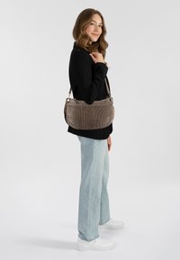 Borsa in tessuto marrone con rifiniture in pelle liscia. Presenta una forma strutturata e arrotondata con tracolla regolabile. Indossata con jeans chiari e sneakers bianche.