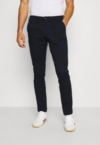 Námořnické modré slim-fit chinos s hladkým povrchem, zapínáním na knoflík a bočními kapsami, spárované s bílými teniskami s červenými a modrými akcenty.