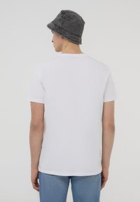 Biała koszulka z krótkim rękawem i okrągłym dekoltem, wykonana z bawełny. Model nosi szary kapelusz typu bucket, pokazany z tylu na jednolitym tle.