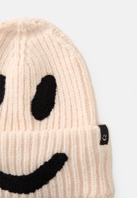 Ljusrosa stickad mössa med ribbat mönster, som har en svart, fluffig smiley-design och en liten logotyp-tagg på sidan.