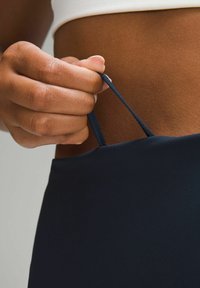 Temno modra športna majica brez rokavov z gladko teksturo, z nastavljivimi naramnicami, ki jih z roko zateguje.