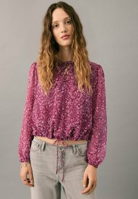 Lila Paisley-Bluse mit langen Ärmeln, gesmoktem Saum mit Kordelzug; halbtransparentes Material; kombiniert mit hellgrauen Jeans.