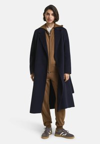 Navigatie lange jas met een geknipte kraag over een bruine hoodie met ritssluiting en bijpassende broek, gecombineerd met grijze sneakers met witte accenten.